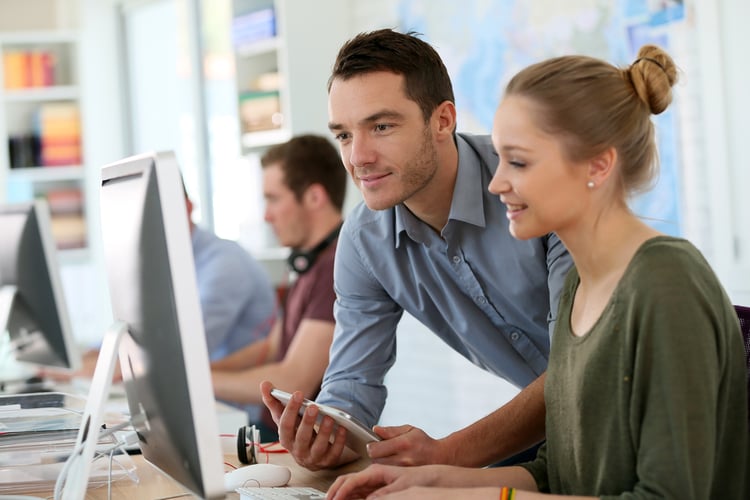Mitarbeterin, die am Computer arbeitet und einem Kollegen mit Tablet den Bildschirm zeigt