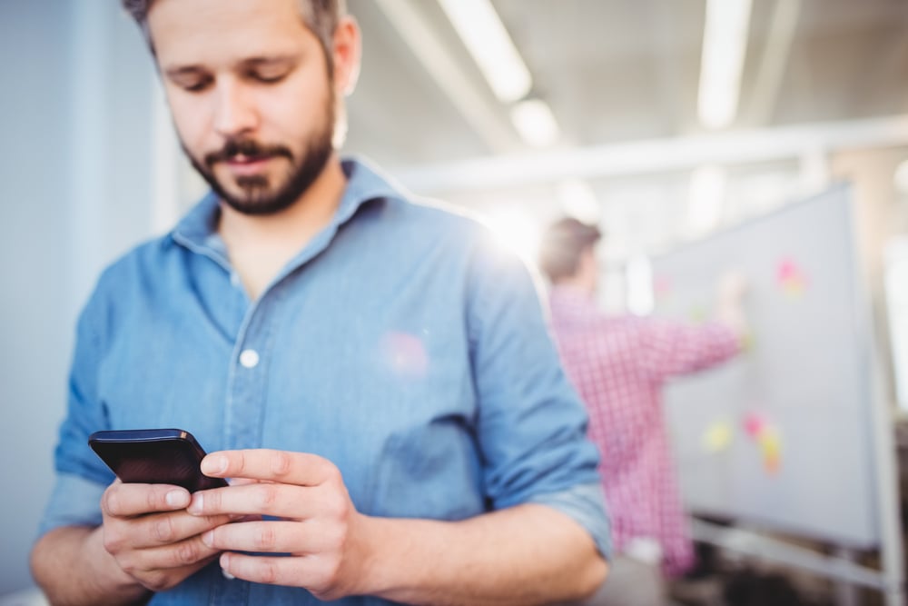 Ein Mitarbeiter beantragt Urlaub am Handy, im Hintergrund arbeitet ein Kollege